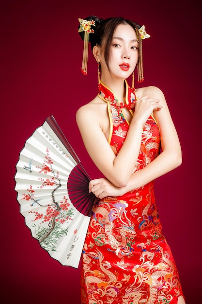 伝統的なチャイナ ドレス qipao ドレスを着てきれいな新鮮な肌を持つ美しい若いアジア女性赤の背景にファンのポーズを保持スタジオ幸せな中国の新年の女性モデルの肖像画