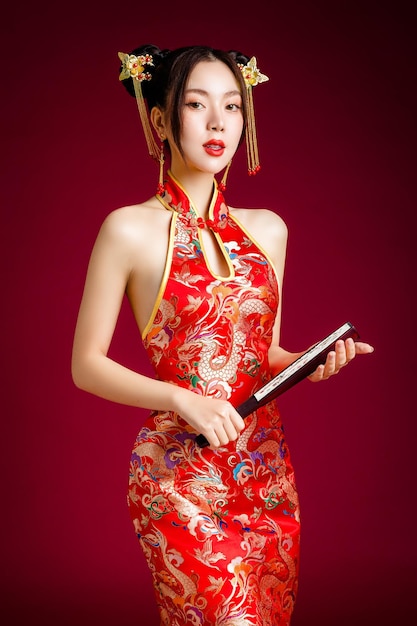 伝統的なチャイナ ドレス qipao ドレスを着てきれいな新鮮な肌を持つ美しい若いアジア女性赤の背景にファンのポーズを保持スタジオ幸せな中国の新年の女性モデルの肖像画