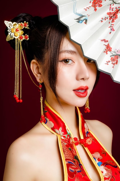 伝統的なチャイナ ドレス qipao ドレスを着てきれいな新鮮な肌を持つ美しい若いアジア女性赤の背景にファンのポーズを保持スタジオ幸せな中国の新年の女性モデルの肖像画