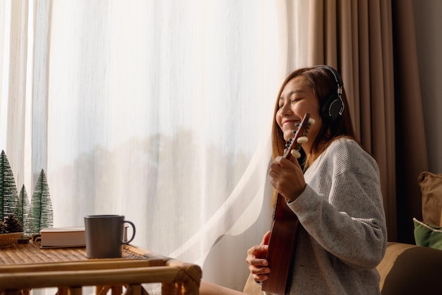 A beautiful young asian woman with headphone enjoy playing an ukulele at home