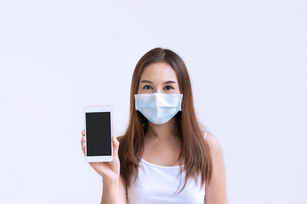 Beautiful young Asian woman with ptotective face mask holding smartphone for copy space on white background