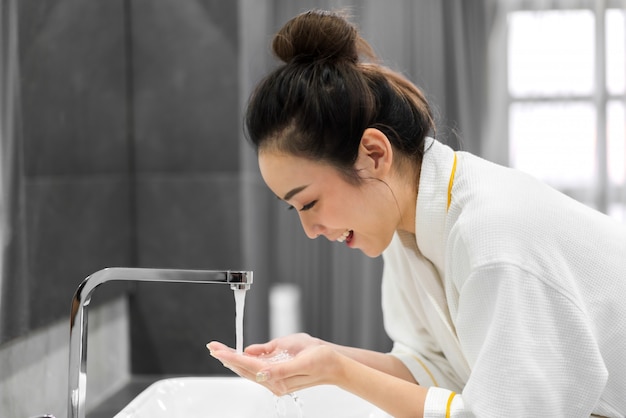 Bella giovane donna asiatica che lava viso pulito con acqua e che sorride davanti allo specchio nel bagno bellezza e stazione termale pelle fresca perfetta