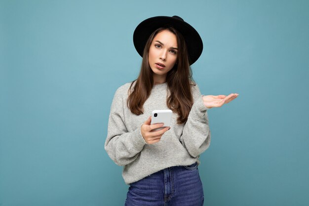 Beautiful young asking dissatisfied brunette woman wearing black hat and grey sweater holding