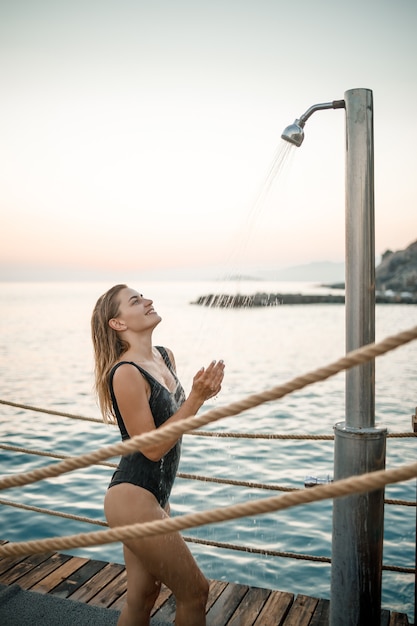 A beautiful young attractive woman in a black swimsuit with a beautiful figure stands under a summer shower on the pier by the sea. She is enjoying her vacation. Selective focus