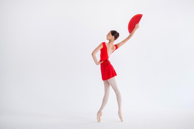 Beautiful young ballerina with a Spanish fan on a white background The ballerina is dressed in a red leotard pink tights pointe shoes