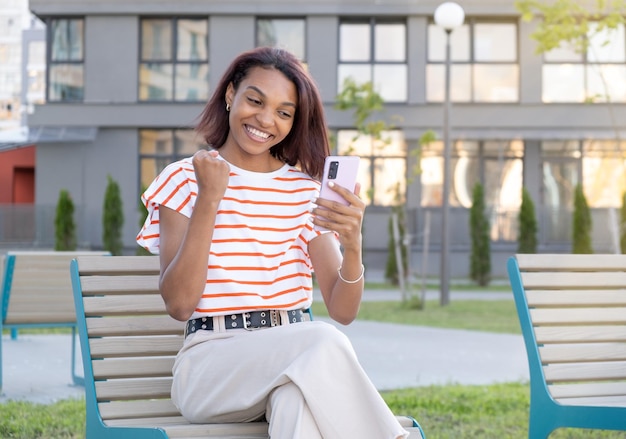 Beautiful young black woman viewing content on her mobile phone feeling happy and excited and celebrating