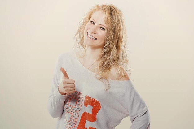 Foto bella giovane donna bionda in jeans e maglietta che mostra un pollice in alto su uno sfondo bianco