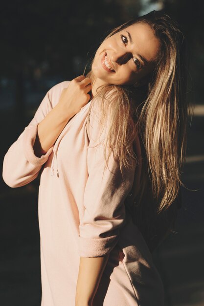 Красивая молодая блондинка женщина в розовой толстовке игриво смотрит на камеру, стоящую в солнечном свете