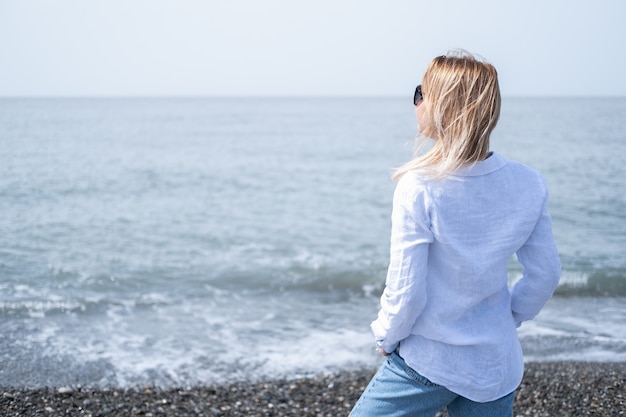 흰 셔츠와 선글라스 바다의 해안에 서 있는 아름 다운 젊은 금발 여자.