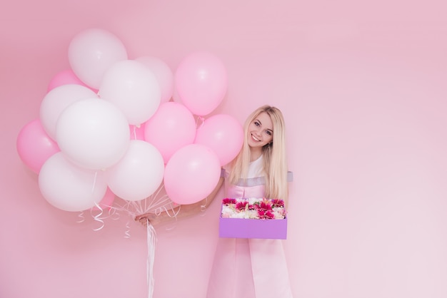 Beautiful young blond woman with balloons and flowers on color wall