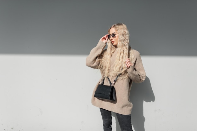 Beautiful young blond woman with curly hairstyle in a fashion trendy beige sweater with a leather bag is wearing vintage sunglasses in the city near a gray wall in the sun light