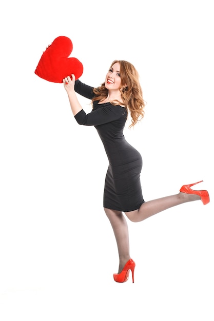 Bella giovane donna bionda con labbra rosse in abito nero e scarpe tacco alto con un cuore rosso nelle mani su uno sfondo bianco, san valentino, trucco di lusso alla moda