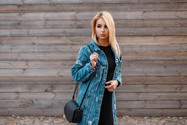 Красивая молодая блондинка со стильной кожаной сумочкой в модной джинсовой куртке в черном платье возле деревянной старинной стены