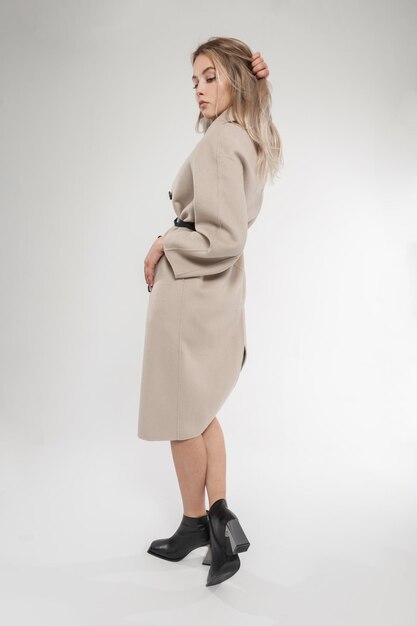 Beautiful young blonde girl in fashionable spring outwear with fashion coat and stylish black leather shoes posing in the studio Womens new collection of shoes