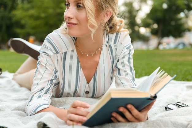 Bella ragazza bionda che si rilassa su un prato al parco, leggendo un libro mentre si sdraia su una coperta