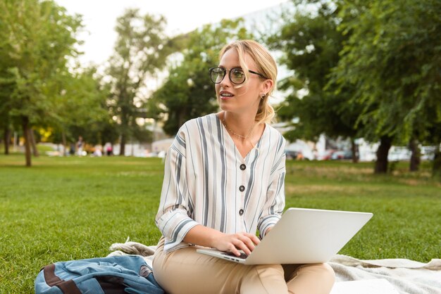 Bella ragazza bionda che si rilassa su un prato al parco, lavora al computer portatile mentre è seduta su una coperta