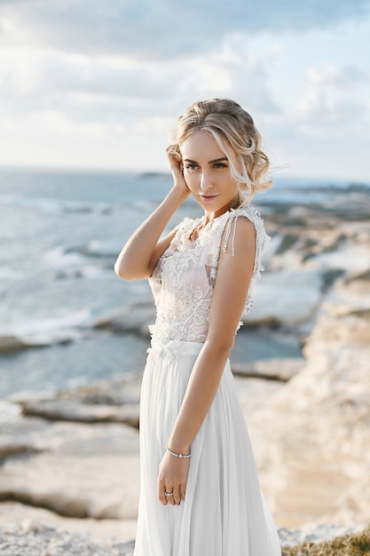 Beautiful young blonde model woman with nude makeup in a fashionable wedding dress walking at the sea coast at Cyprus