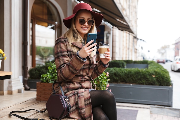 Beautiful young blonde stylish woman wearing a coat relaxing outdoors, taking a selfie