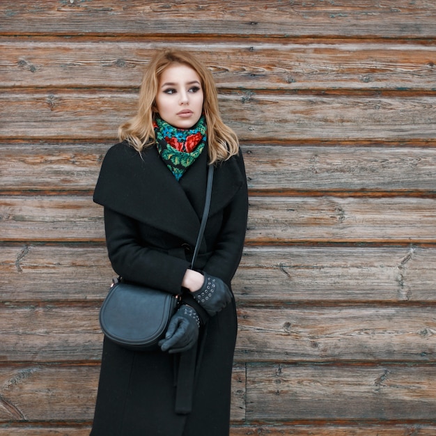 黒いコートと木製の壁の近くのスタイリッシュなハンドバッグの美しい若いブロンドの女性