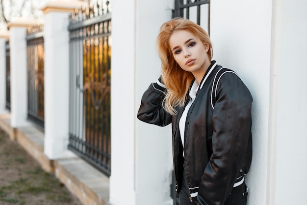 Beautiful young blonde woman in a black summer stylish jacket in black jeans in a white polo shirt standing near a vintage metal fence. Fashionable modern girl.