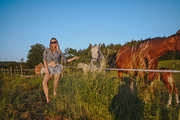 Beautiful young blonde woman dressed safari style in hat and plaid shirt posing with thoroughbreds