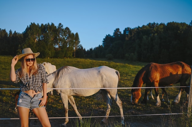 Beautiful young blonde woman dressed safari style in hat and plaid shirt posing with thoroughbreds