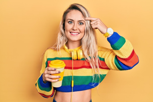 Beautiful young blonde woman drinking cup of coffee wearing headphones smiling pointing to head with one finger, great idea or thought, good memory