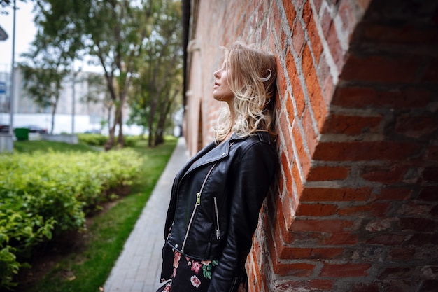 A beautiful young blonde woman in a leather black jacket, a pretty short dress and a hat walks in the Zaryadye Park, Moscow. 