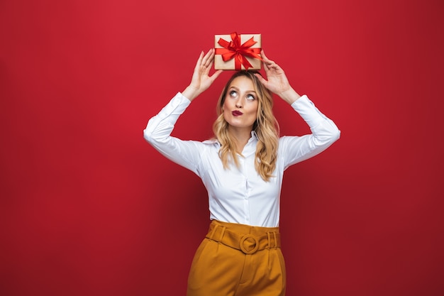 Фото Красивая молодая блондинка женщина, стоящая изолированно на красном фоне, держа подарочную коробку на голове
