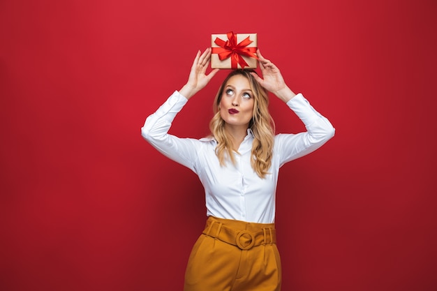 Beautiful young blonde woman standing isolated over red background, holding gift box on her head