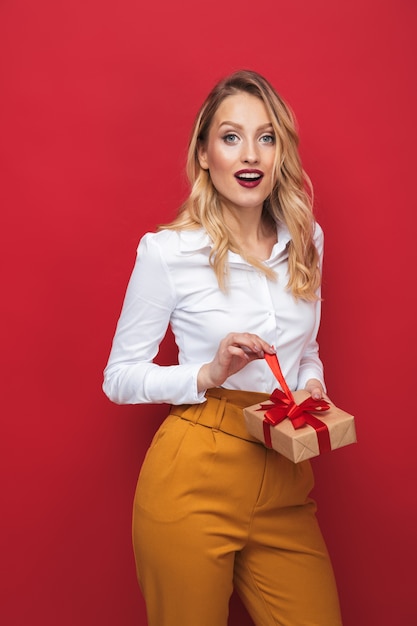 Beautiful young blonde woman standing isolated over red background, holding gift box