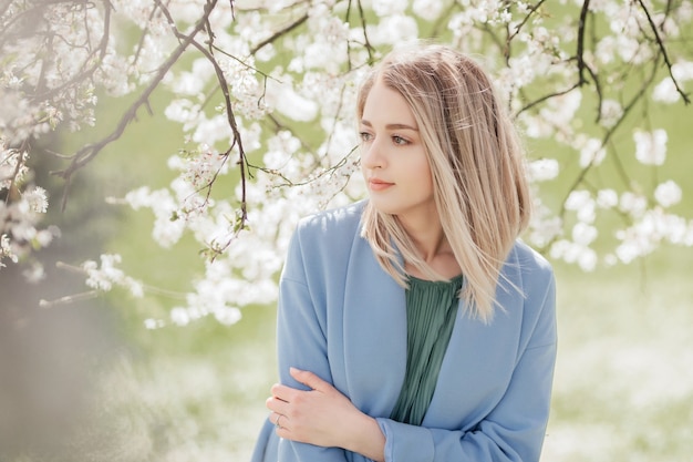 Красивая молодая блондинка стоит возле яблони в зеленом платье и синем пальто