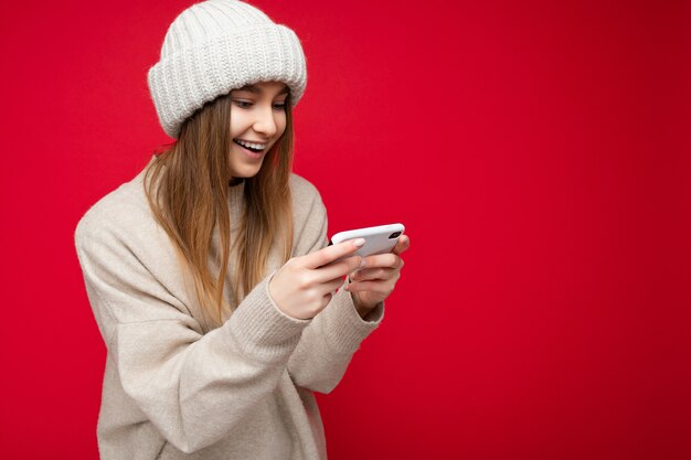 Beautiful young blonde woman wearing casual beige sweater and beige hat isolated over red background