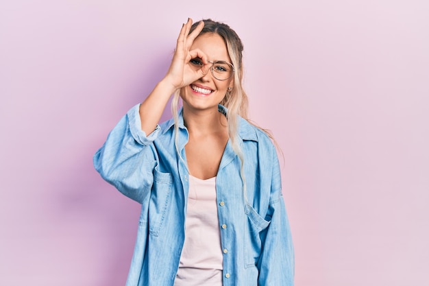 Beautiful young blonde woman wearing casual clothes and glasses doing ok gesture with hand smiling eye looking through fingers with happy face