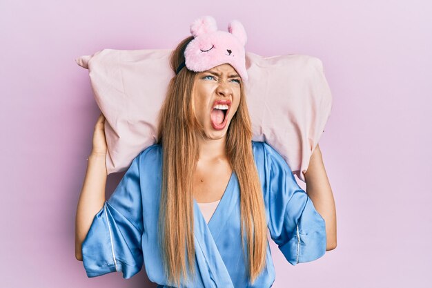 Beautiful young blonde woman wearing sleep mask and pajama sleeping on pillow angry and mad screaming frustrated and furious shouting with anger looking up