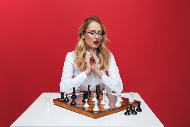 Beautiful young blonde woman wearing white shirt sitting at the table, playing chess isolated over red background