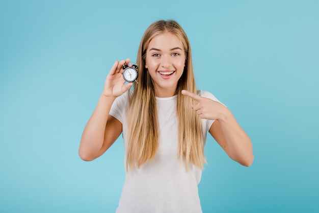 Beautiful young blonde woman with alarm clock showing time isolated over blue