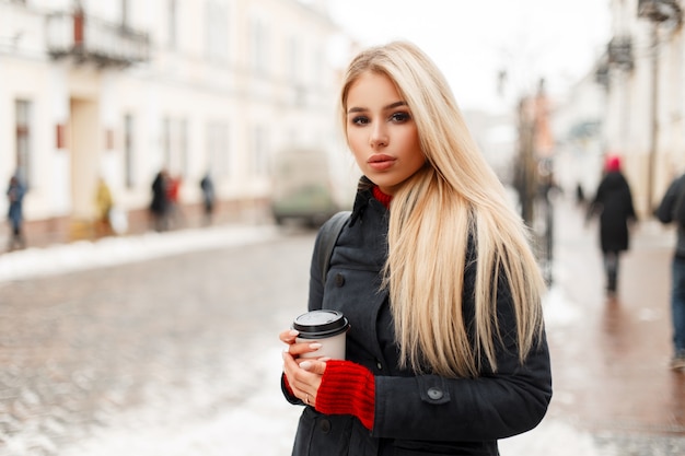 Bella giovane donna bionda con caffè in un cappotto invernale di moda in città