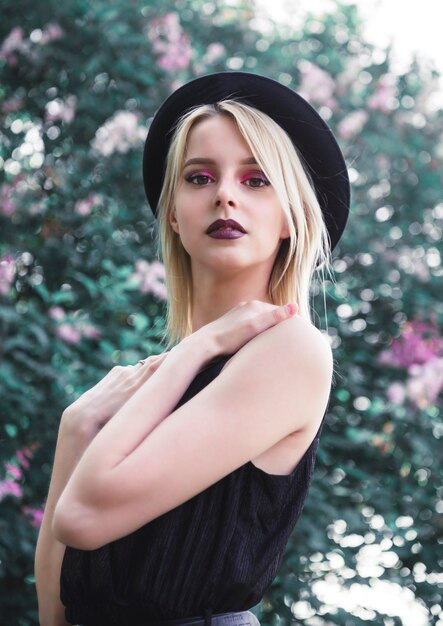 Beautiful young blonde woman with pink makeup in a black top and hat against a surface of blooming lilac