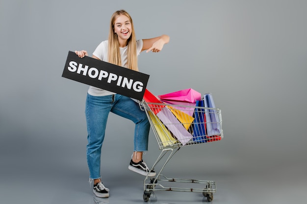 Beautiful young blonde woman with shopping sign and pushcart with colorful shopping bags isolated over grey