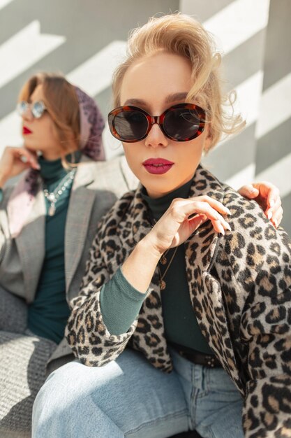 Beautiful young blonde woman with vintage sunglasses in leopard jacket and sweater with jeans on the street in sunny day Urban retro female style Two pretty stylish girls