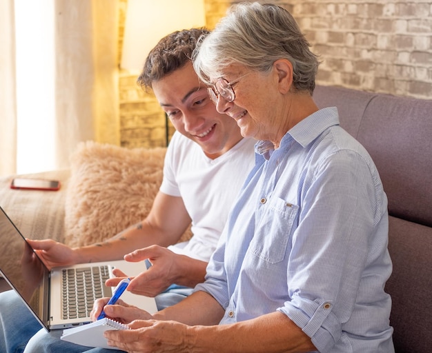 Beautiful young boy sitting on sofa at home helping senior\
grandmother surf with laptop on the web while she takes notes\
younger generations care for older relatives are teaching computers\
to use