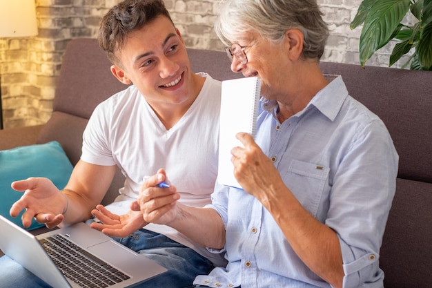 Beautiful young boy sitting on sofa at home helping senior\
grandmother surf with laptop on the web while she takes notes\
younger generations care for older relatives teaching how to use\
computer