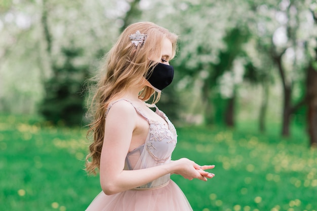 Beautiful young bride in a wedding dress and a black medical mask on her face near a blooming apple trees. Covid-19 protection.