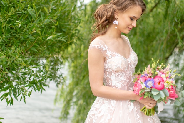 Beautiful young bride in white wedding dress posing at nature with bouquet of flowers.
