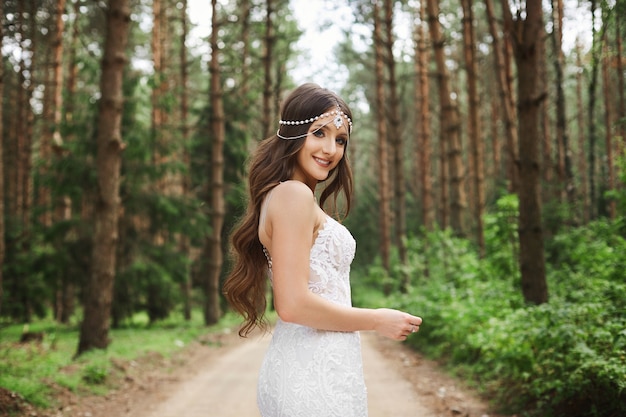 Beautiful young bride with wedding hairstyle with jewelry in lace dress holding a bouquet of flowers in her hands and posing at the forest in the early morning