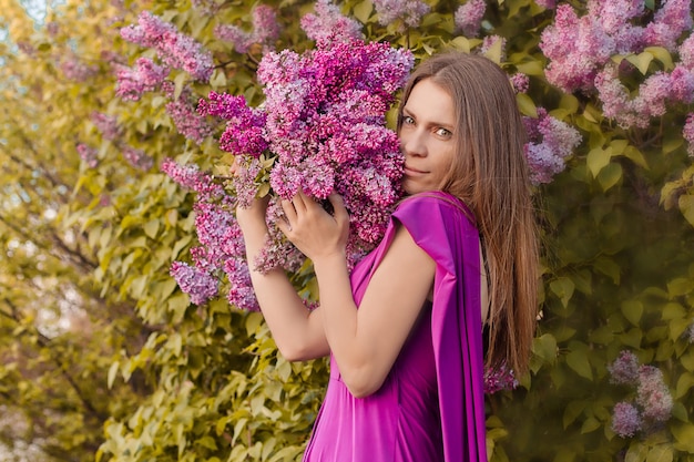 a beautiful young brownhaired girl in a pink dress stands against the background of a lilac bush