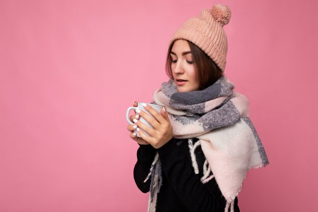 Beautiful young brunet woman wearing black sweater hat and warm scarf isolated over pink wall holding paper mug  drinking hot tea and looking down. copy space