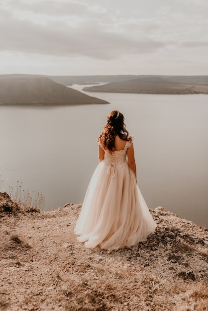 Beautiful young brunette bride in a white wedding dress with a crown on her head stands on a cliff against the background of the river and islands