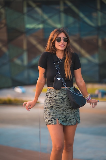 Beautiful young brunette caucasian businesswoman outside the background office building. With green skirt and sunglasses walking